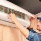 A young male contractor wearing a blue cap checks an air conditioner in a bare room.