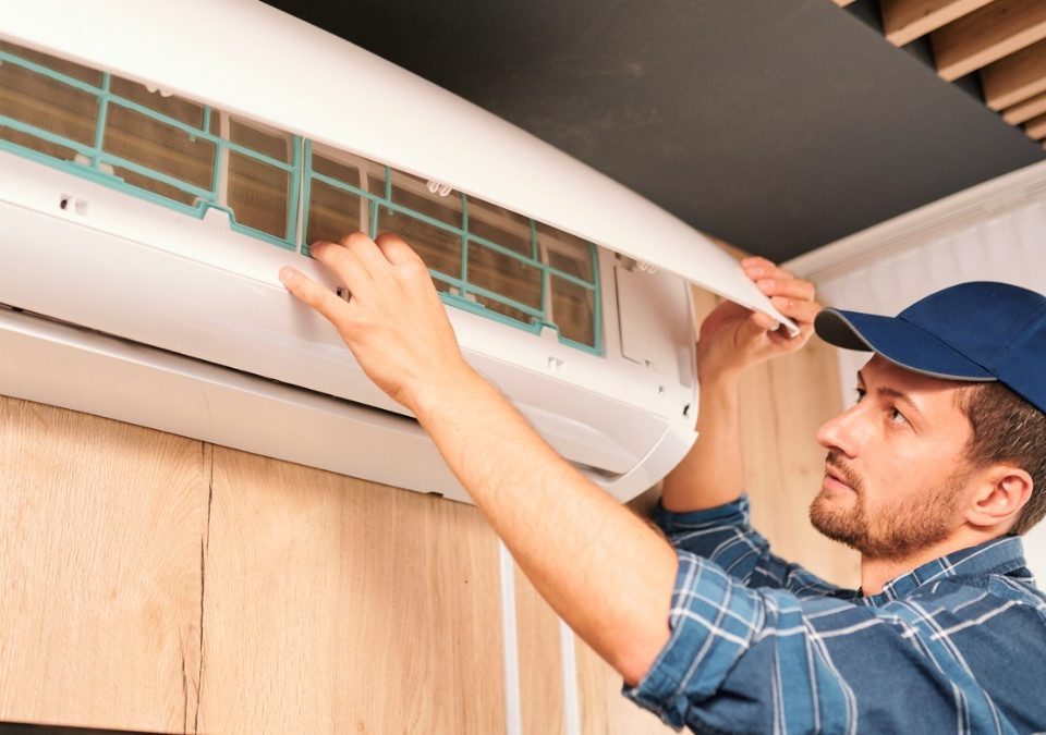 A young male contractor wearing a blue cap checks an air conditioner in a bare room.