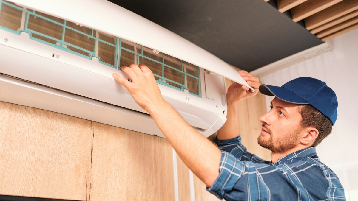 A young male contractor wearing a blue cap checks an air conditioner in a bare room.