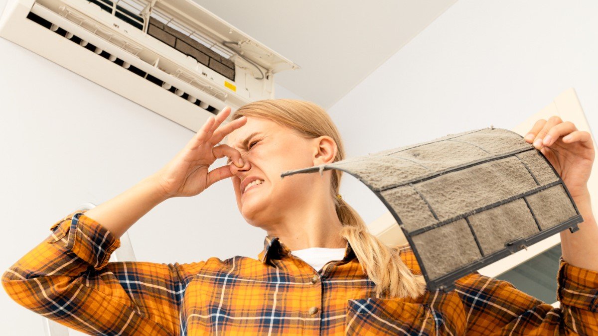 A female contractor in an orange shirt pinches her nose because of a moldy AC filter.