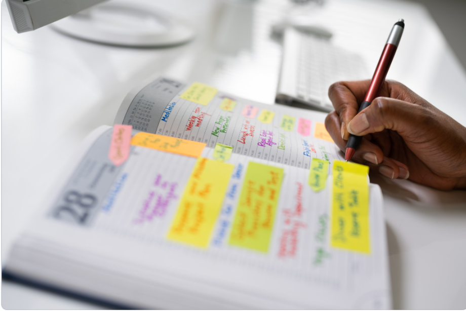 Close-up of a person writing in a planner filled with colourful sticky notes and reminders, with a computer keyboard in the background.