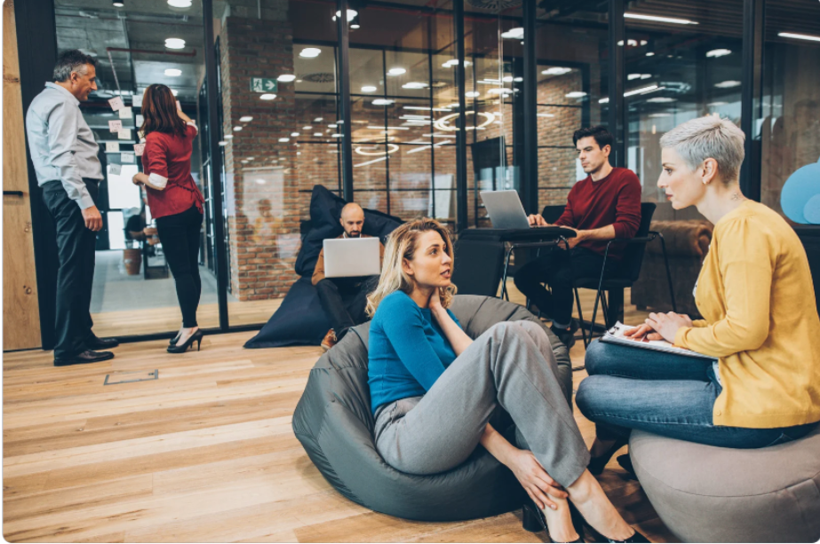 Group of colleagues having a casual meeting in a modern office space, with some sitting on bean bags and others working on laptops.