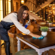 Smiling female restaurant staff member cleaning a table with a blue cloth and sanitizer in a warmly lit restaurant interior.