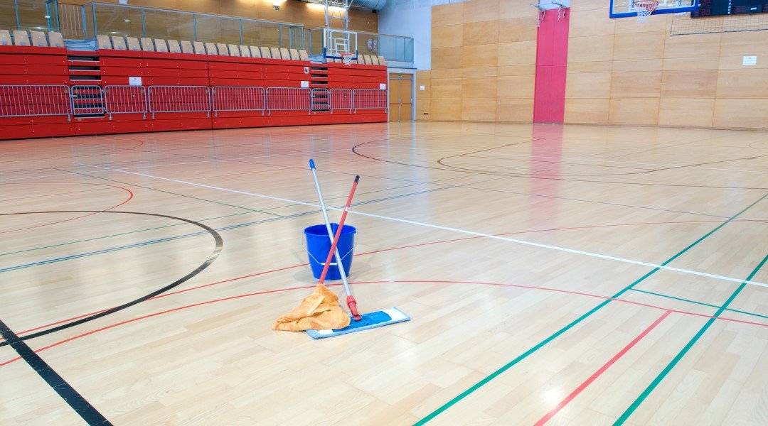 A freshly mopped gymnasium floor with a blue bucket and mop in the foreground, highlighting the cleanliness and readiness for sports activities.