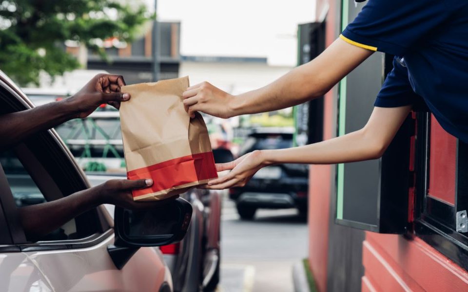 A fast-food worker hands a paper bag of food to a customer in a car at a drive-thru window, illustrating a convenient meal pickup process.