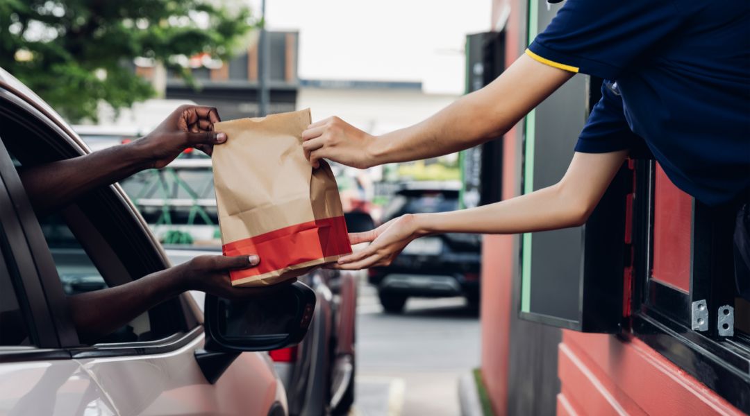 A fast-food worker hands a paper bag of food to a customer in a car at a drive-thru window, illustrating a convenient meal pickup process.