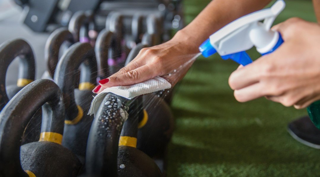 A person cleaning kettlebells with a spray bottle and cloth in a gym, ensuring hygiene and safety for gym users.