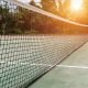 A close-up view of a tennis court net at sunrise, with the sun casting a warm glow on the court and surrounding trees in the background.