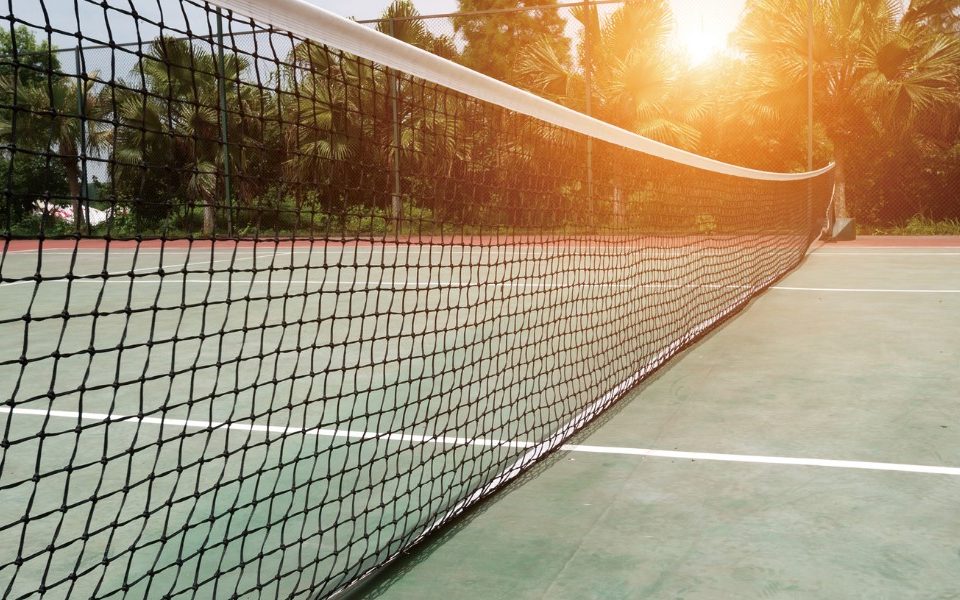 A close-up view of a tennis court net at sunrise, with the sun casting a warm glow on the court and surrounding trees in the background.