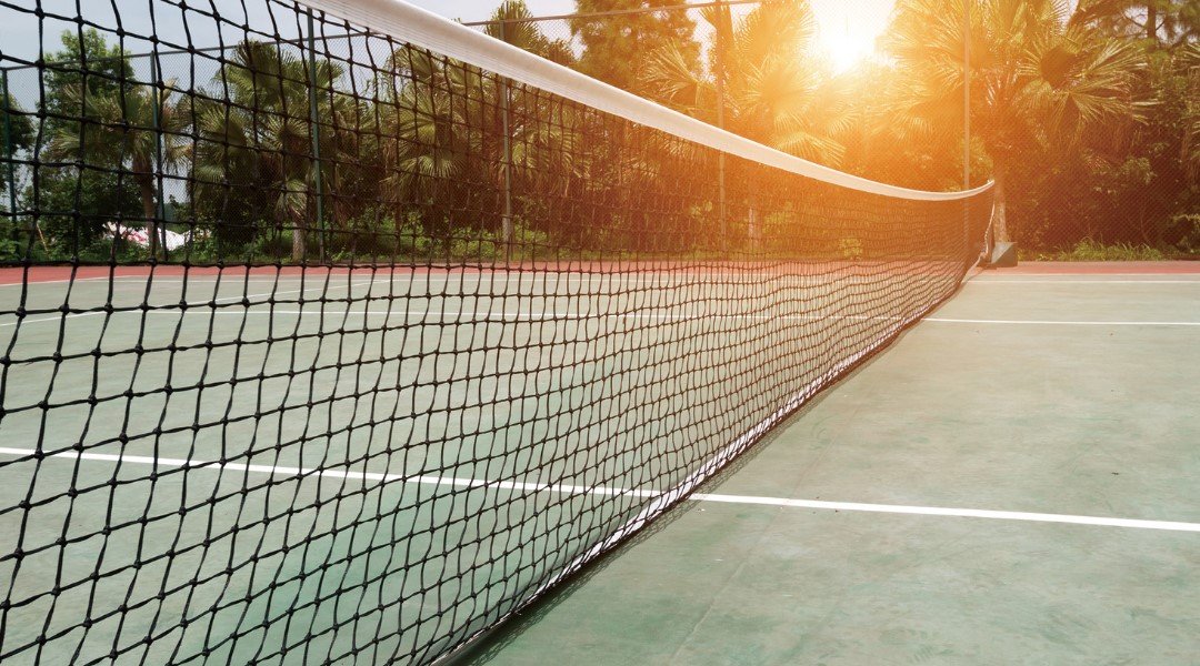 A close-up view of a tennis court net at sunrise, with the sun casting a warm glow on the court and surrounding trees in the background.