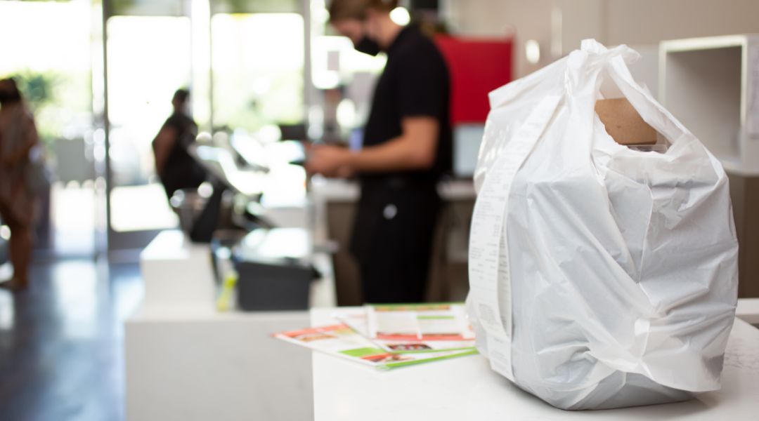 A takeout food order in a white plastic bag placed on a counter, ready for customer pickup, with a blurred background of a restaurant interior.