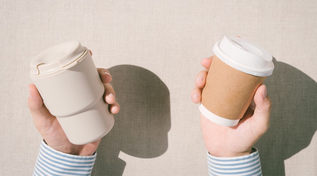 Two hands holding different coffee cups against a neutral fabric background.