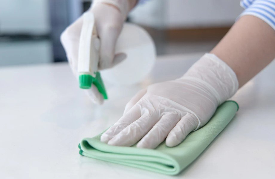 A person wearing gloves using a spray bottle and microfiber cloth to clean a surface, demonstrating effective and hygienic cleaning practices. 