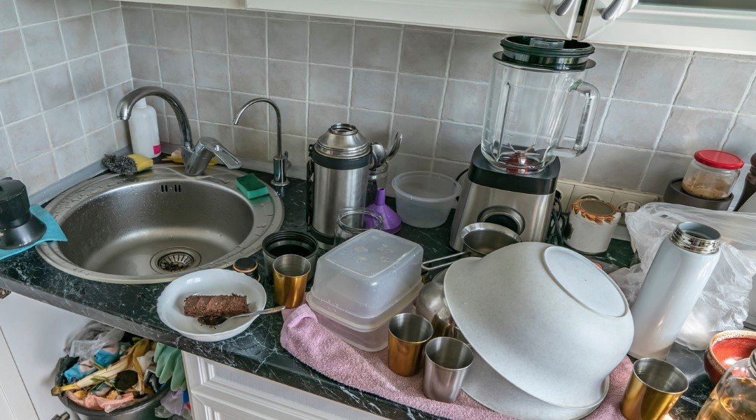 A kitchen counter filled with dirty dishes, a blender, cups, and utensils, showing a busy or unclean kitchen environment.