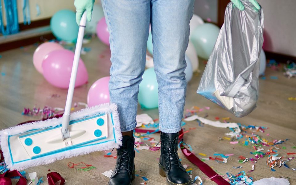 Person wearing gloves, holding a trash bag, and using a mop to clean up confetti and balloons after a party in a room.