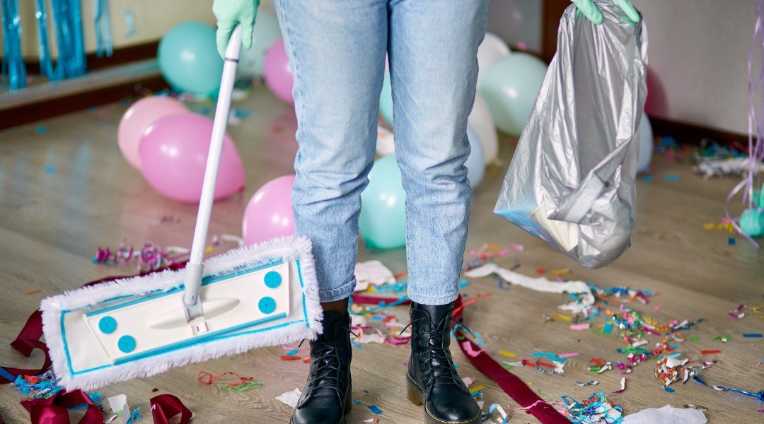 Person wearing gloves, holding a trash bag, and using a mop to clean up confetti and balloons after a party in a room.