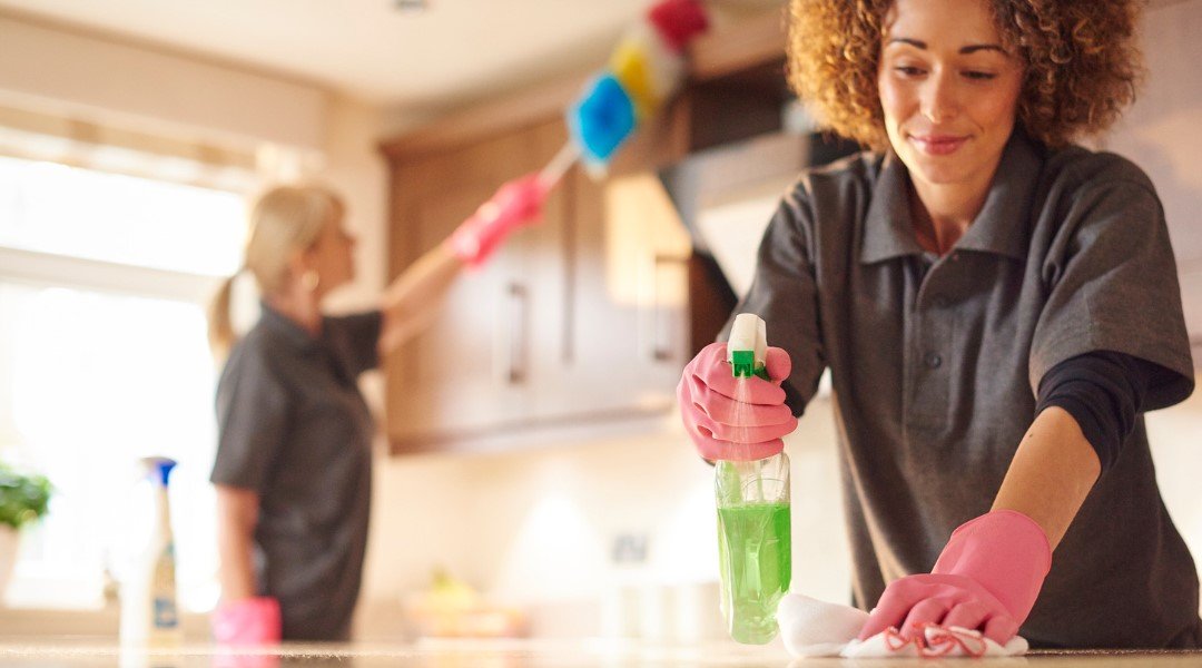 Two professional cleaners in uniforms and gloves thoroughly clean a kitchen, using sprays and dusters to ensure a spotless finish.