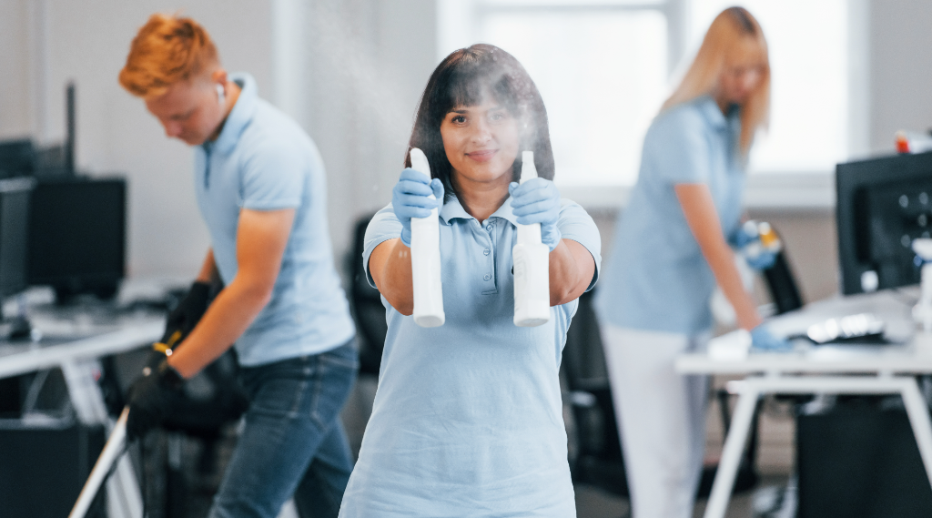 A cleaning team working in an office. A woman sprays disinfectant toward the camera, while other workers clean desks in the background.