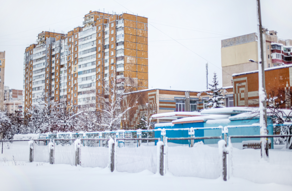 A snowy city scene with tall brick apartment buildings, frosted trees, and snow-covered fences, showcasing a typical winter day.