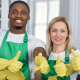 Four cheerful cleaning staff in green aprons and yellow gloves pose in a bright kitchen, giving a thumbs-up gesture, symbolizing professionalism and teamwork.